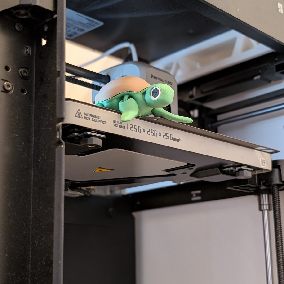 A small green plush turtle toy with a tan shell sits on the print bed of a black 3D printer.