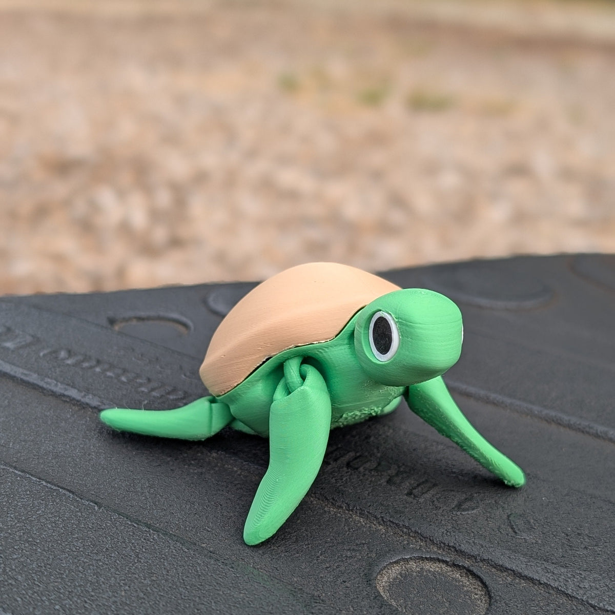 A small turtle-shaped decorative figure with a green body and tan shell, placed on a dark textured surface outdoors with a blurred gravel and foliage background.